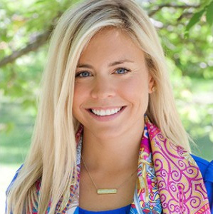 blonde woman in blue top and pink scarf smiling outside.