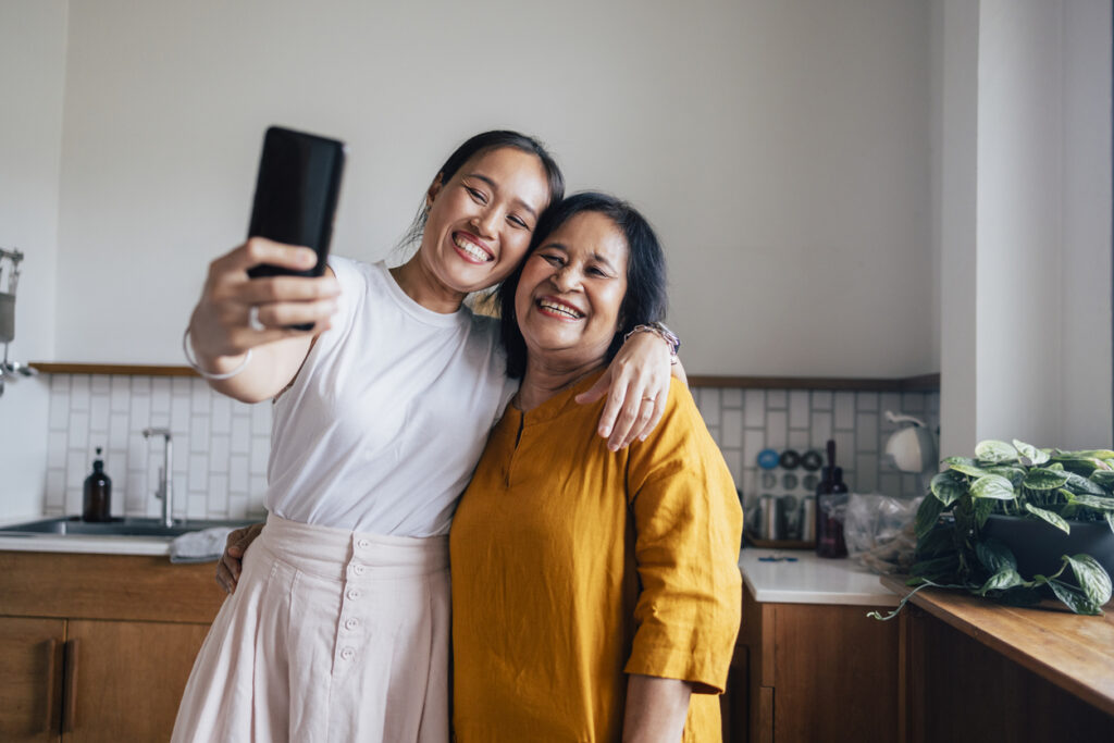 Asian daughter and her mother taking a selfie with a smartphone in their kitchen.